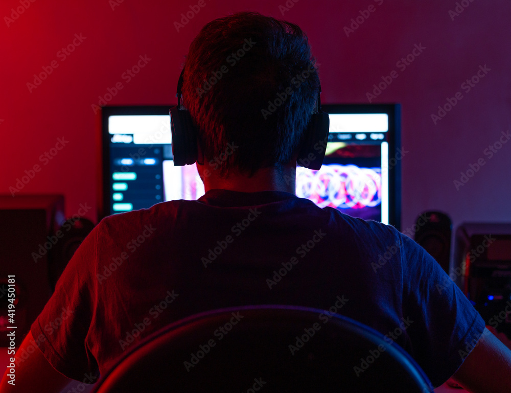 Back view of calm young brunet in casual wear and wireless headset browsing contemporary computer with blank screen while sitting at table in dark room Back view of calm young brunet in casual wear and wireless headset browsing contemporary computer with blank screen while sitting at table in dark room