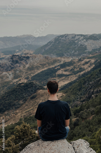 Back view of unrecognizable male hiker sitting on rocky slope of mountain and observing landscape while travelling through highlands