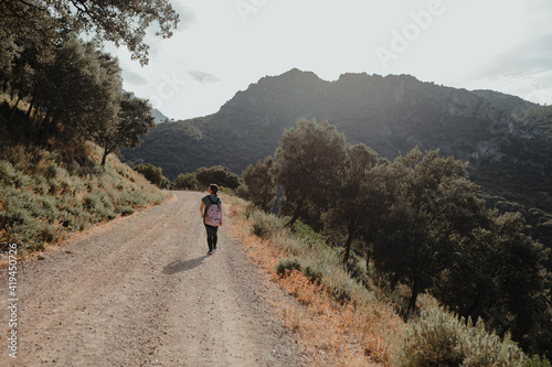 Full body back view of unrecognizable female hiker with backpack walking on narrow road towards rocky mountain peak surrounded by green forest during travel in Spain photo