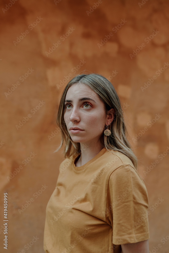 Young content blonde female in casual wear with wavy hair looking away while standing on brown background in town Young content blonde female in casual wear with wavy hair looking away while standing on brown background in town