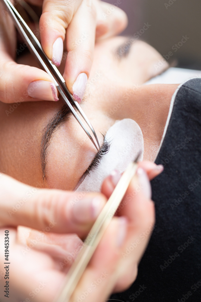 Crop professional female master with tweezer applying artificial eyelashes on young female client in face mask in light modern salon Crop professional female master with tweezer applying artificial eyelashes on young female client in face mask in light modern salon