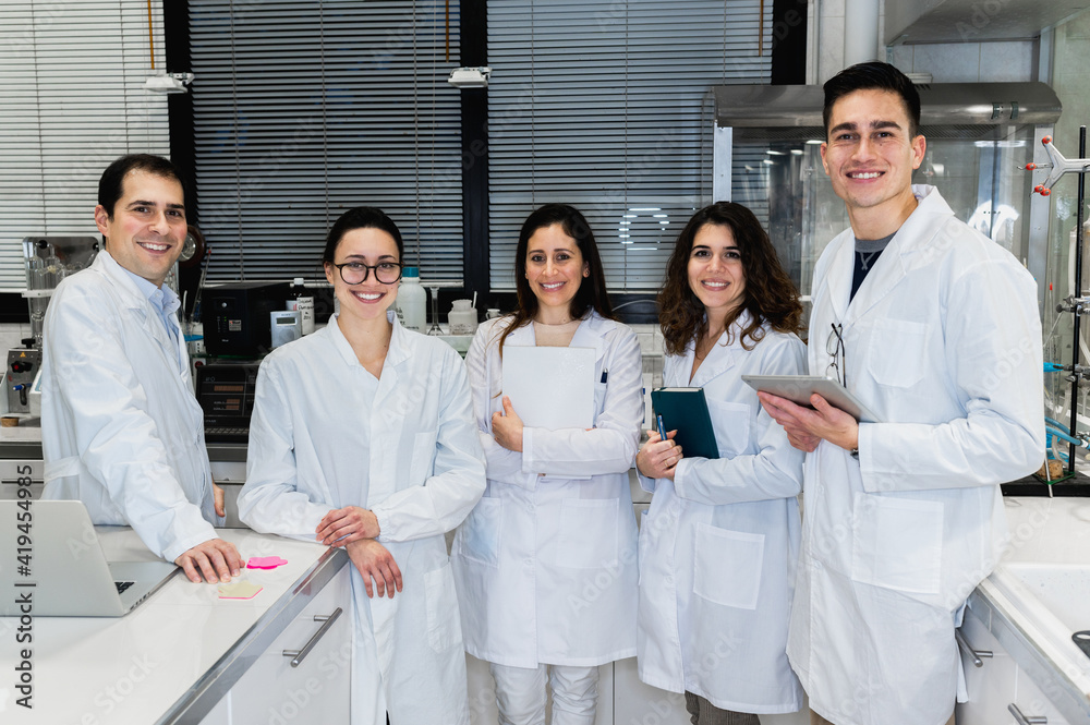 Group of positive young scientific researchers in white coats smiling and looking at camera while standing together in modern lab Group of positive young scientific researchers in white coats smiling and looking at camera while standing together in modern lab
