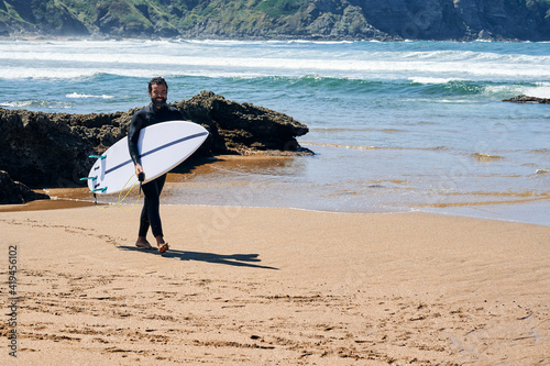 Adult man in wetsuit with surfboard smiling happily standing on seashore against hills photo