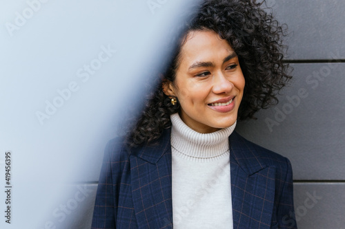 Delighted self assured young ethnic female in stylish clothes looking away while standing against gray striped wall