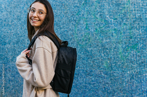 Side view crop female student in casual wear with backpack standing against blue tiled wall and looking away with smile photo