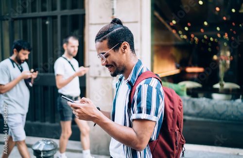 Positive dark skinned african american hipster guy holding mobile phone for checking notification walking on street, smiling african american male tourist exploring city on journey browsing map