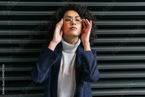 Serious self assured young ethnic female in stylish clothes touching eyeglasses and looking away while standing against gray striped wall