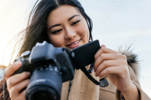 Ethnic young happy Asian female photographer shooting photo on professional photo camera on city street