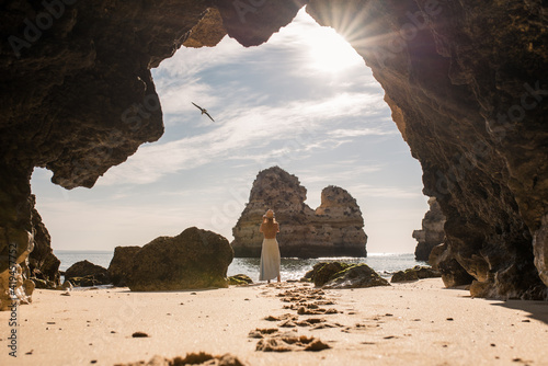Back view of unrecognizable female in casual clothes and hat standing on sand in entrance of cave near sea looking at a bird in Algarve, Portugal