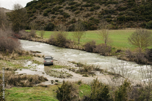 Modern SUV parked on shore of rapid river flowing in hilly terrain covered with plants photo