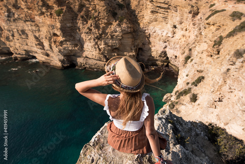 Back view of unrecognizable young woman in summer clothes and straw hat sitting in cliff admiring landscape at Cape Saint Vincent in Sagres, portugal