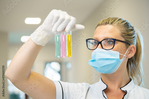 Serious female scientist in mask and latex gloves standing with glass test tubes filled with chemical liquids of various colors and working in lab photo