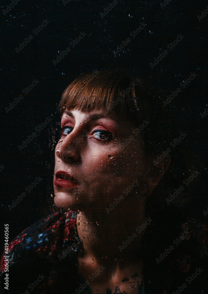 Unemotional female with ear piercing standing behind translucent glass with water droplets looking at camera Unemotional female with ear piercing standing behind translucent glass with water droplets looking at camera
