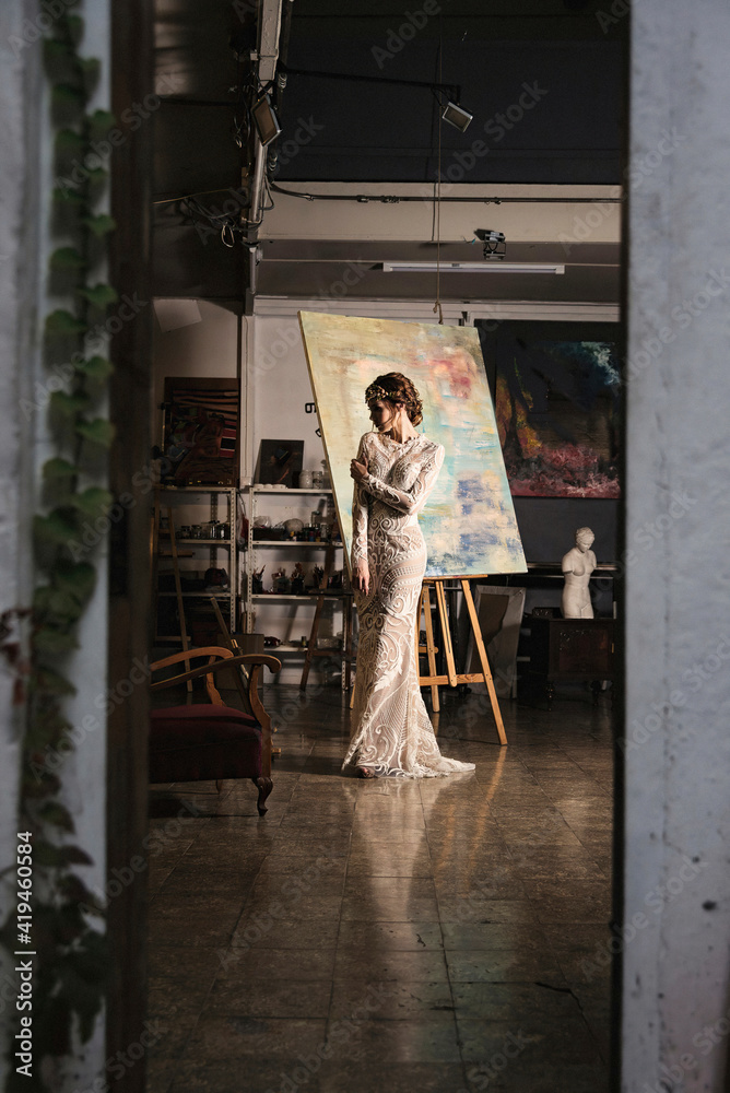 Full body of charming young slender female in elegant maxi bridal gown standing near easel with painting in dark art studio Full body of charming young slender female in elegant maxi bridal gown standing near easel with painting in dark art studio