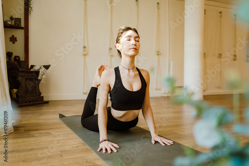 Young concentrated female in sportswear demonstrating Raja Bhujangasana pose with closed eyes on yoga mat in house