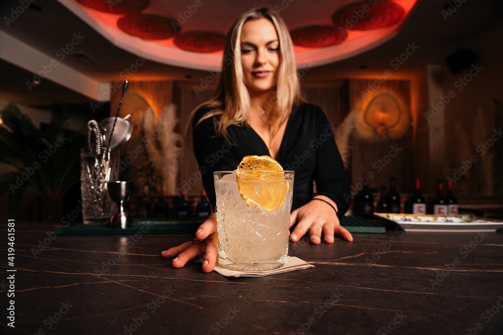 © Gustavo Pozo/ADDICTIVE STOCK - Content young female bartender with long blond hair serving cold alcohol cocktail decorated with lemon slice in stylish restaurant