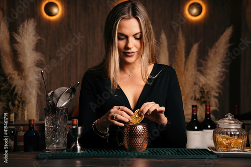 Self assured focused young female barkeeper with long blond hair in stylish outfit decorated cocktail with lemon slices while standing at counted in stylish bar