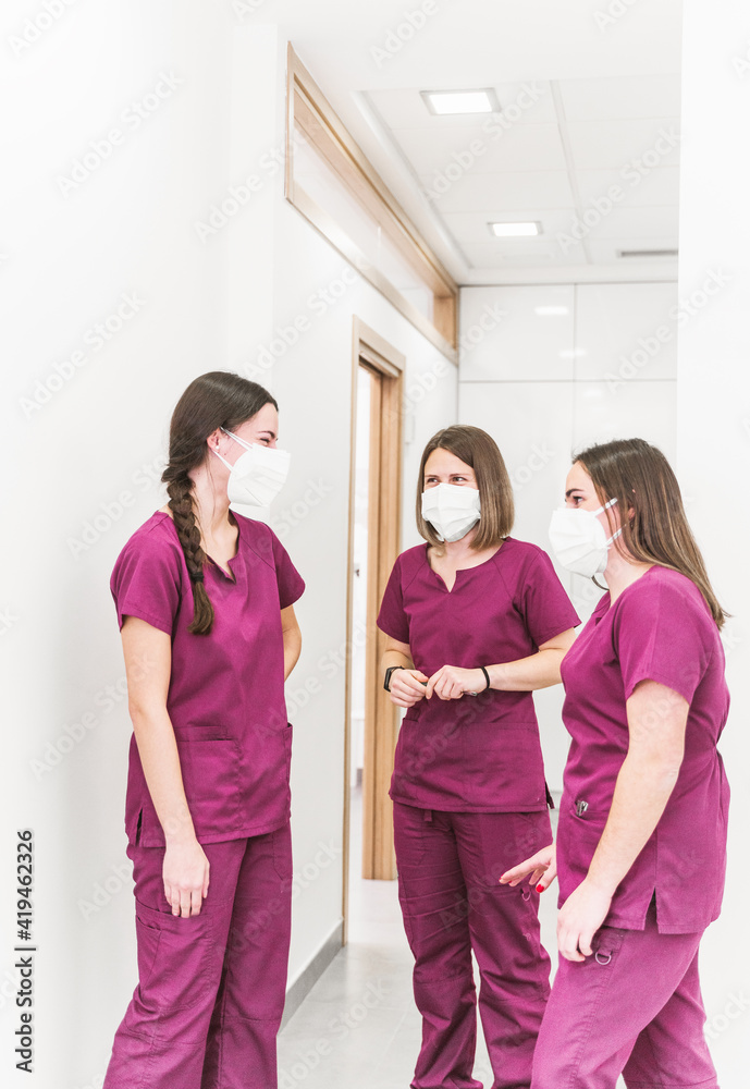 Group of young content nurses in medical uniform and masks having conversation in modern hospital corridor Group of young content nurses in medical uniform and masks having conversation in modern hospital corridor