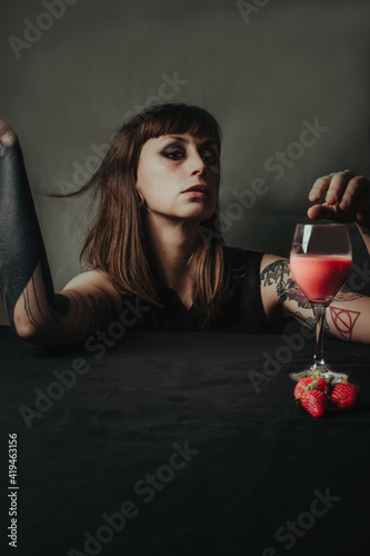 Young female with dark makeup and raised arms against transparent glass of sweet refreshing drink with fresh strawberries photo