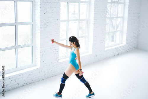 Fototapeta Naklejka Na Ścianę i Meble -  Caucasian female in bodysuit training lower body during pilates workout with dumbbells equipment, slim woman in knee pads slimming during aerobic practice for keeping healthy lifestyle and wellness