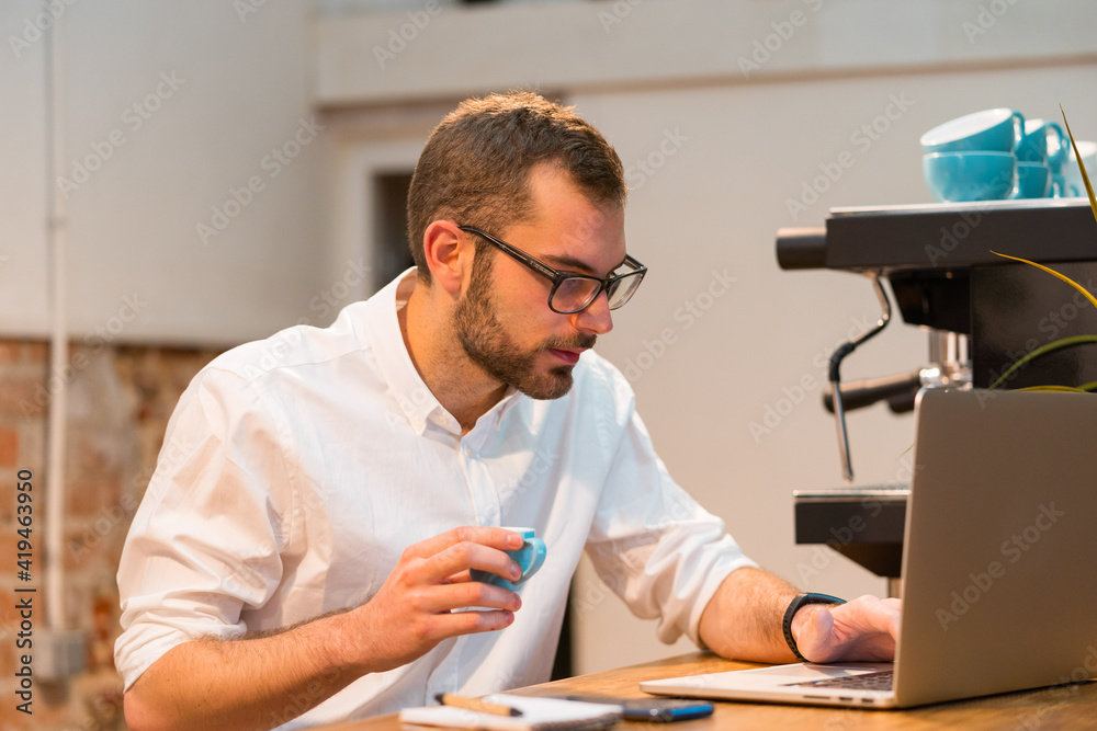 Focused male barista sitting at wooden counter with laptop and taking notes in notebook while working in coffee shop Focused male barista sitting at wooden counter with laptop and taking notes in notebook while working in coffee shop