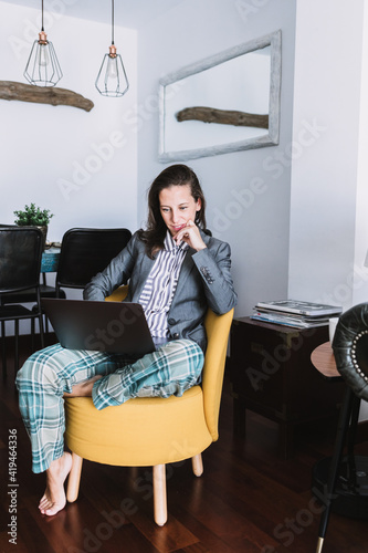 Full body of young female remote employee wearing formal blazer and homey pajama pants sitting on chair and working on laptop at home