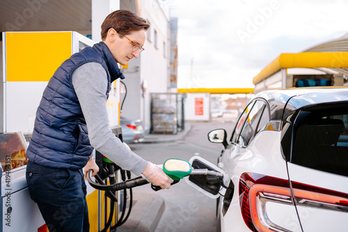 Side view of serious young man in casual clothes and eyeglasses with plastic gloves filling up fuel into modern car in petrol station in daytime photo