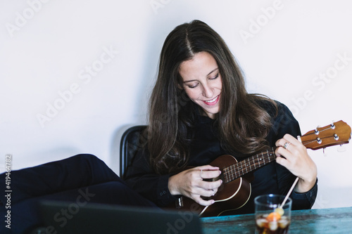 Happy young female looking at laptop screen and playing ukulele while having online lesson at shabby wooden table at home
