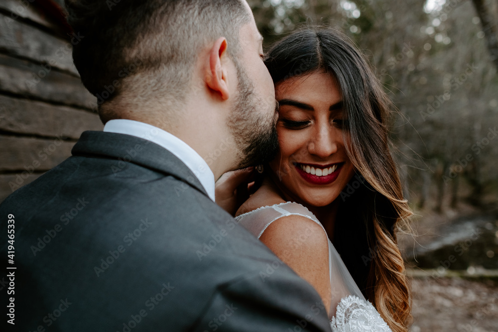 Side view of young groom in elegant suit kissing smiling ethnic bride while standing in nature on wedding day Side view of young groom in elegant suit kissing smiling ethnic bride while standing in nature on wedding day