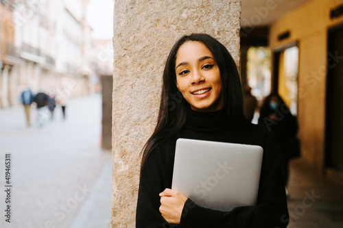 Charming young ethnic female with netbook smiling at camera while standing at stone column on city street photo