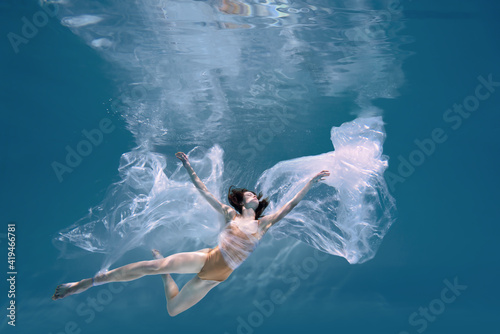 Fashionable and athletic girl free diver alone in the depths of the ocean. Swimmer brunette diving deep in ocean on blue underwater background. Pollution, plastic and ecology concept