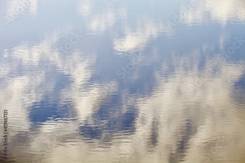 White clouds and blue sky reflected on surface of calm blue water. Beautiful glowing ripples on water surface close-up. Highlights and reflections on water surface with abstract pattern close-up. © ST-art