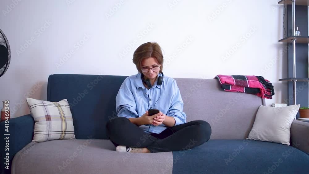 A woman in glasses with headphones is typing text in a mobile phone while sitting on the couch.