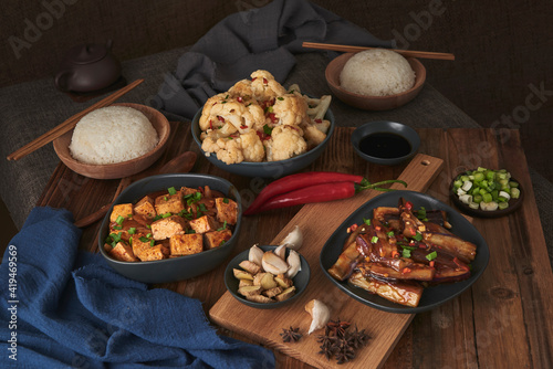 Mala tofu and yuxiang, chinese vegan dishes, accompanied by a bowl of rice, cauliflower, soy sauce and a Japanese teapot on top of a wooden table decorated with fabrics photo