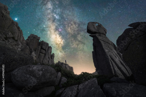 Silhouette of man standing on hill with big ricks among green plants with colorful night sky with stars and milky way on background