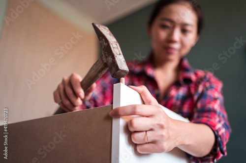 Focused ethnic female using hammer for nailing wooden dowels while assembling new furniture in apartment photo