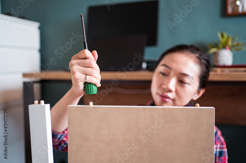 Ethnic Asian female sitting on floor thumbing wooden board with screws while assembling new furniture at home