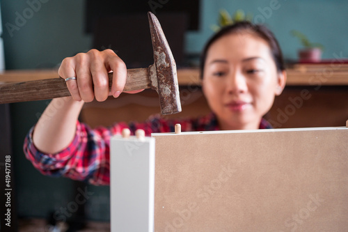 Focused ethnic female using hammer for nailing wooden dowels while assembling new furniture in apartment photo