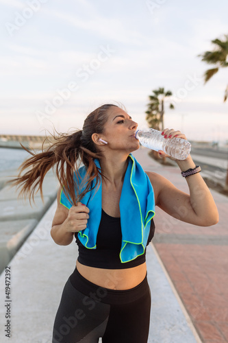 Young fit female jogger with towel on neck drinking water from bottle while having break after running on urban waterfront photo