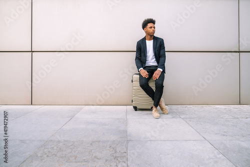 Peaceful black male traveler sitting on suitcase near building in airport while waiting for departure and looking away photo