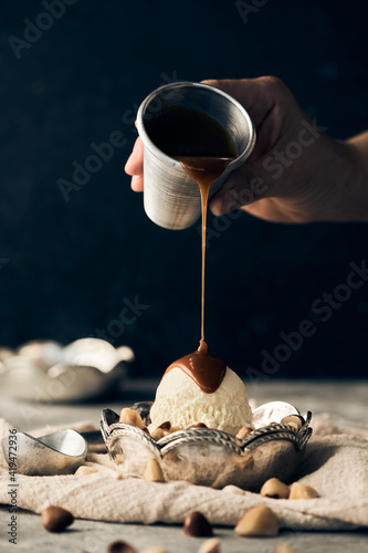 Unrecognizable crop person pouring sweet caramel topping on vanilla ice cream scoop in bowl placed on table with scattered nuts