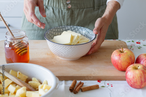 Unrecognizable person mixing butter with flour in bowl while preparing dough for crumble near butter and apples with cinnamon on table at home