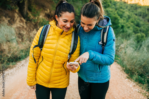 Cheerful female hikers with backpacks and in warm jackers standing on road in nature and orientating on GPS map on smartphone during trekking photo