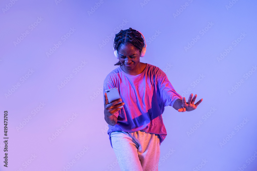 Serious African American teenage girl with smartphone in headphones standing in studio with bright glowing neon lights Serious African American teenage girl with smartphone in headphones standing in studio with bright glowing neon lights