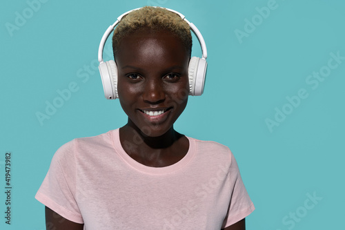 Serene beautiful African American female smiling looking at camera while listening to music in headphones against blue background
