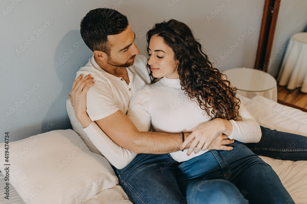 Loving couple sitting on soft bed in bedroom and tenderly hugging while enjoying weekend together at home Loving couple sitting on soft bed in bedroom and tenderly hugging while enjoying weekend together at home