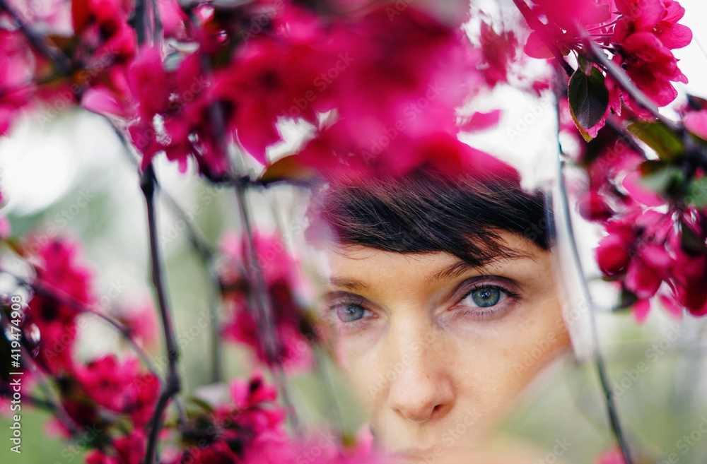 Crop pensive adult female with short hair recreating in green garden near bright blossoming flowers and looking at camera