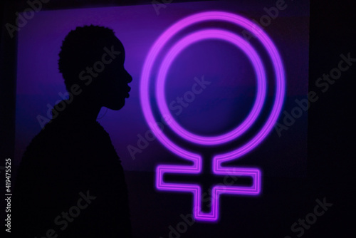 Side view of unrecognizable trendy black model silhouette standing near neon female symbol glowing with purple light