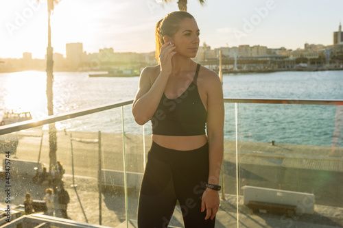 Positive fit female in sportswear adjusting wireless earbuds while preparing for outdoor fitness training on city quay at sunset time
