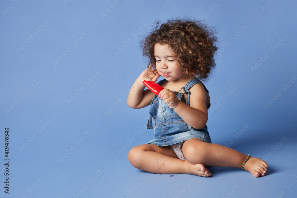 Full body of delighted little girl sitting with melting popsicle against blue background Full body of delighted little girl sitting with melting popsicle against blue background